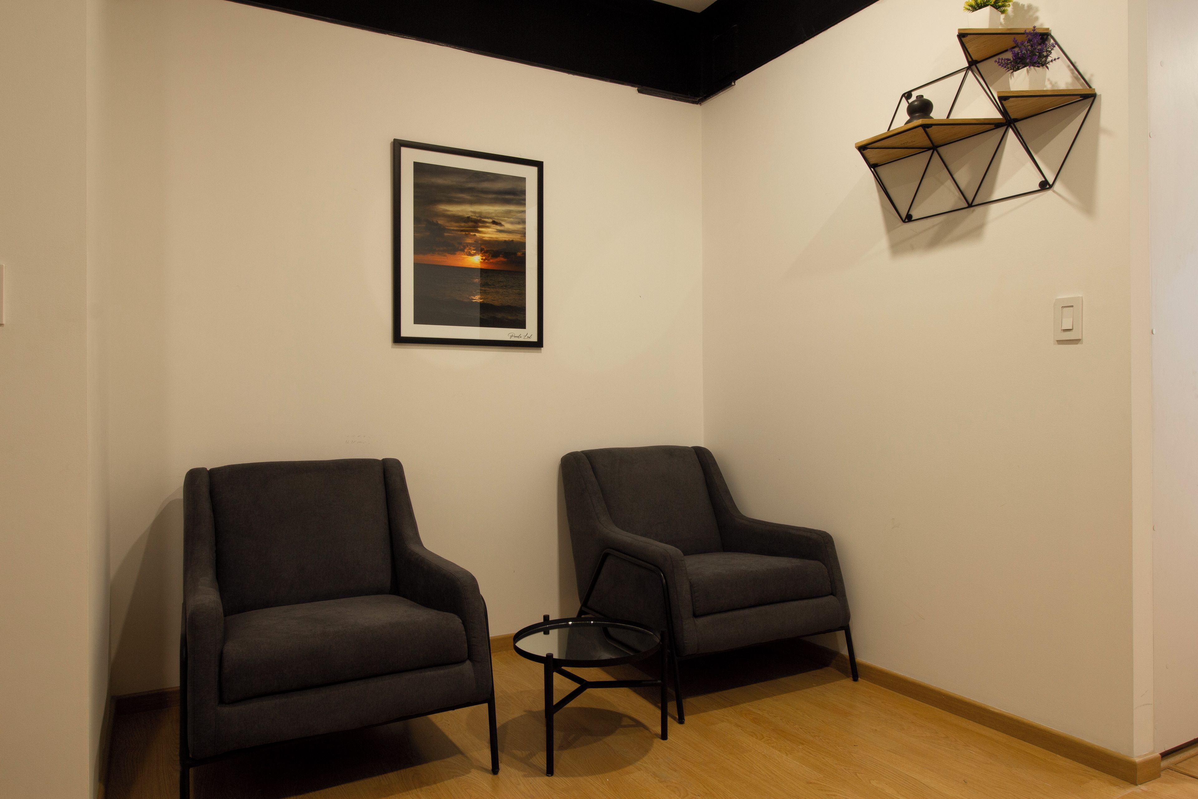 Una habitación con dos sillones oscuros, una mesita redonda pequeña, un cuadro enmarcado de un atardecer y una estantería geométrica con objetos decorativos.