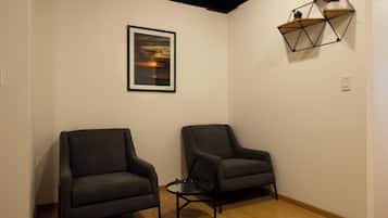 Una habitación con dos sillones oscuros, una mesita redonda pequeña, un cuadro enmarcado de un atardecer y una estantería geométrica con objetos decorativos.