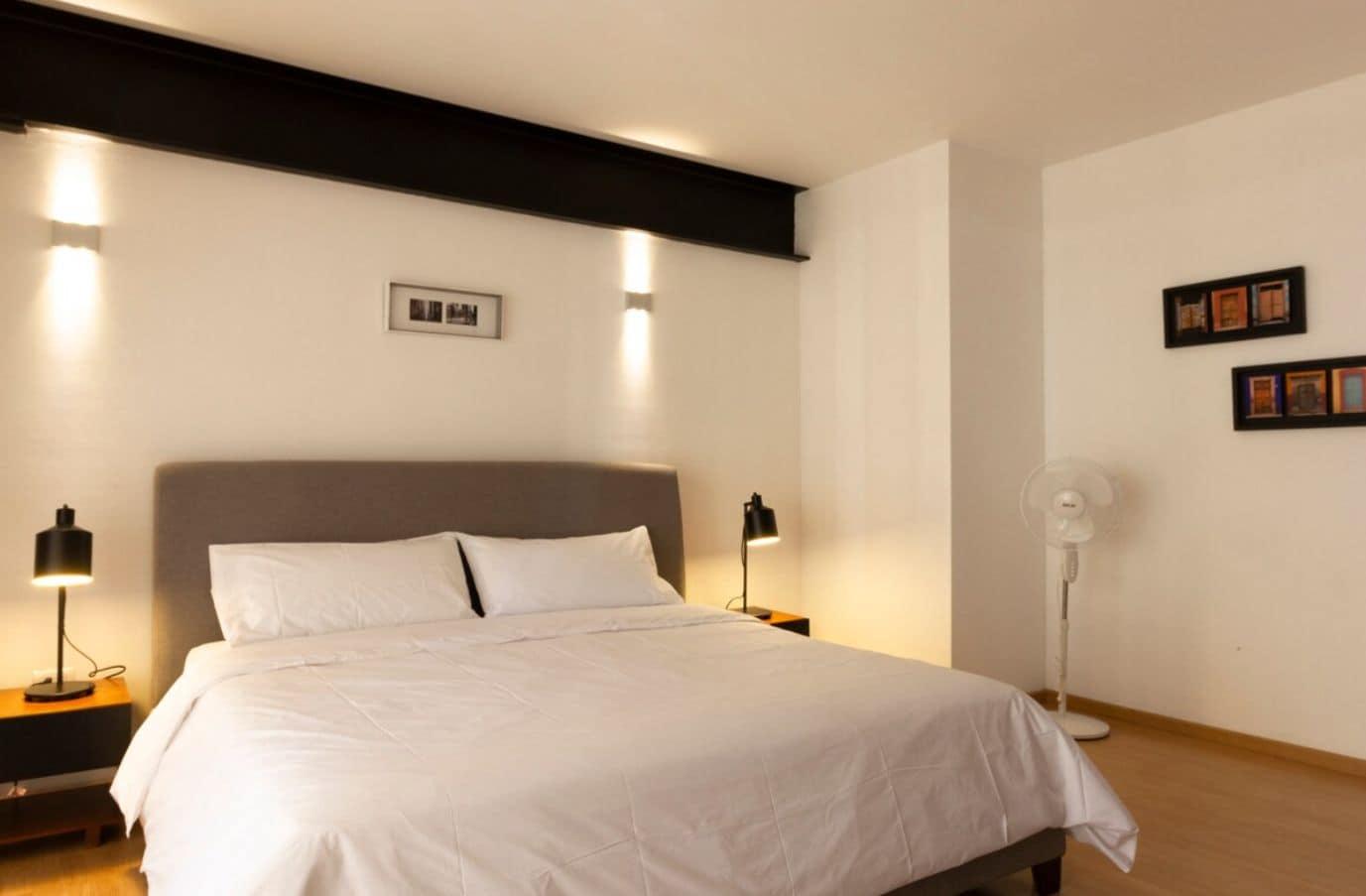 Una cama bien hecha en una habitación de hotel, con un cabecero, dos mesitas de noche con lámparas, un ventilador de pie y cuadros enmarcados en la pared.