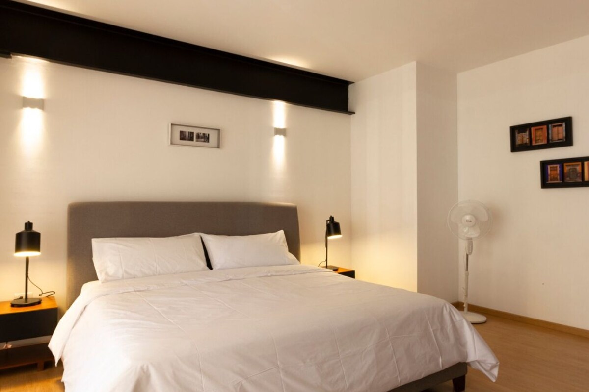 A neatly made bed in a hotel room with a headboard, two bedside tables with lamps, a standing fan, and framed pictures on the wall.