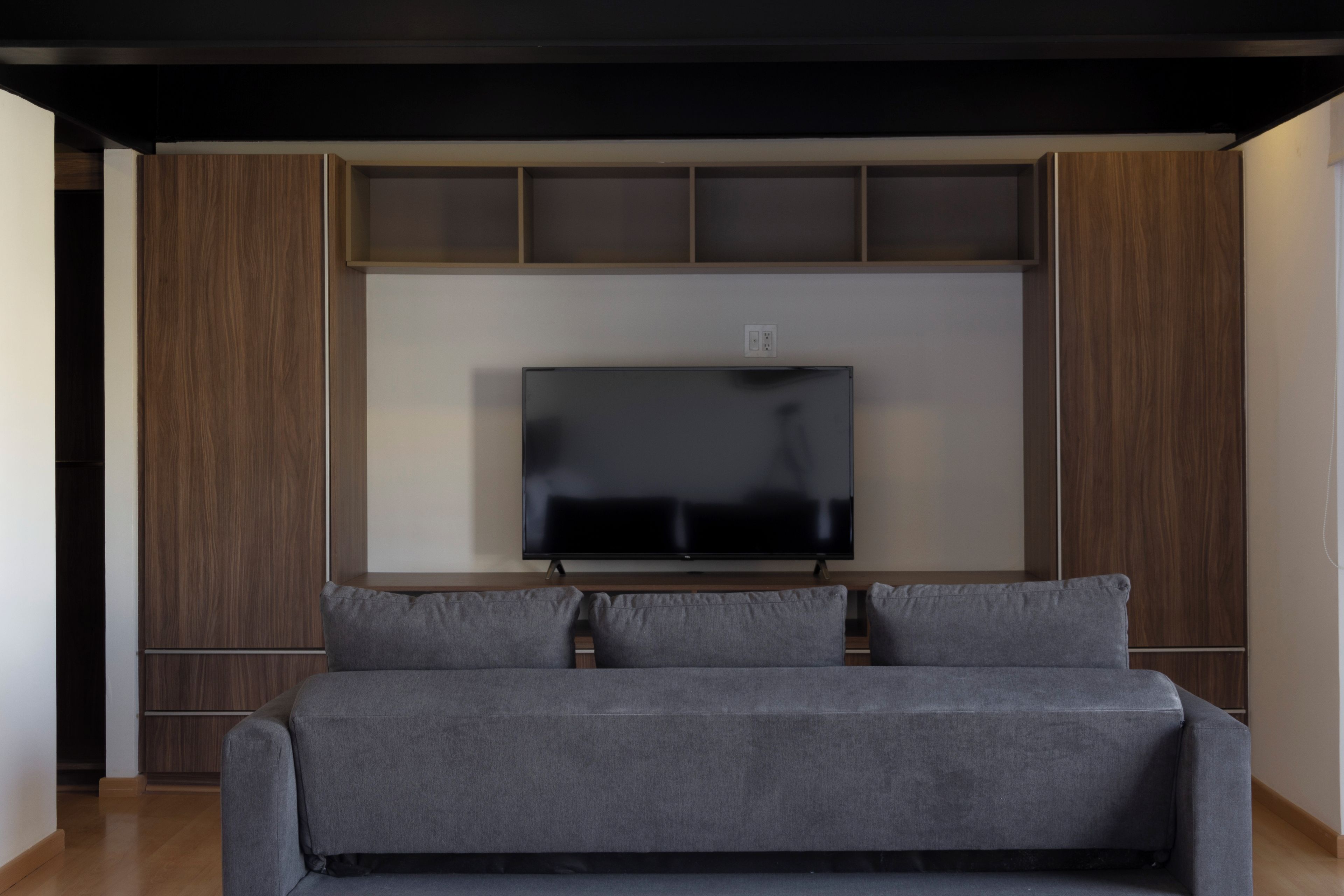 Ein modernes Wohnzimmer mit einem großen Flachbildfernseher an einer Holzwand, einem grauen Sofa und eingebauten Regalen über dem Fernseher.