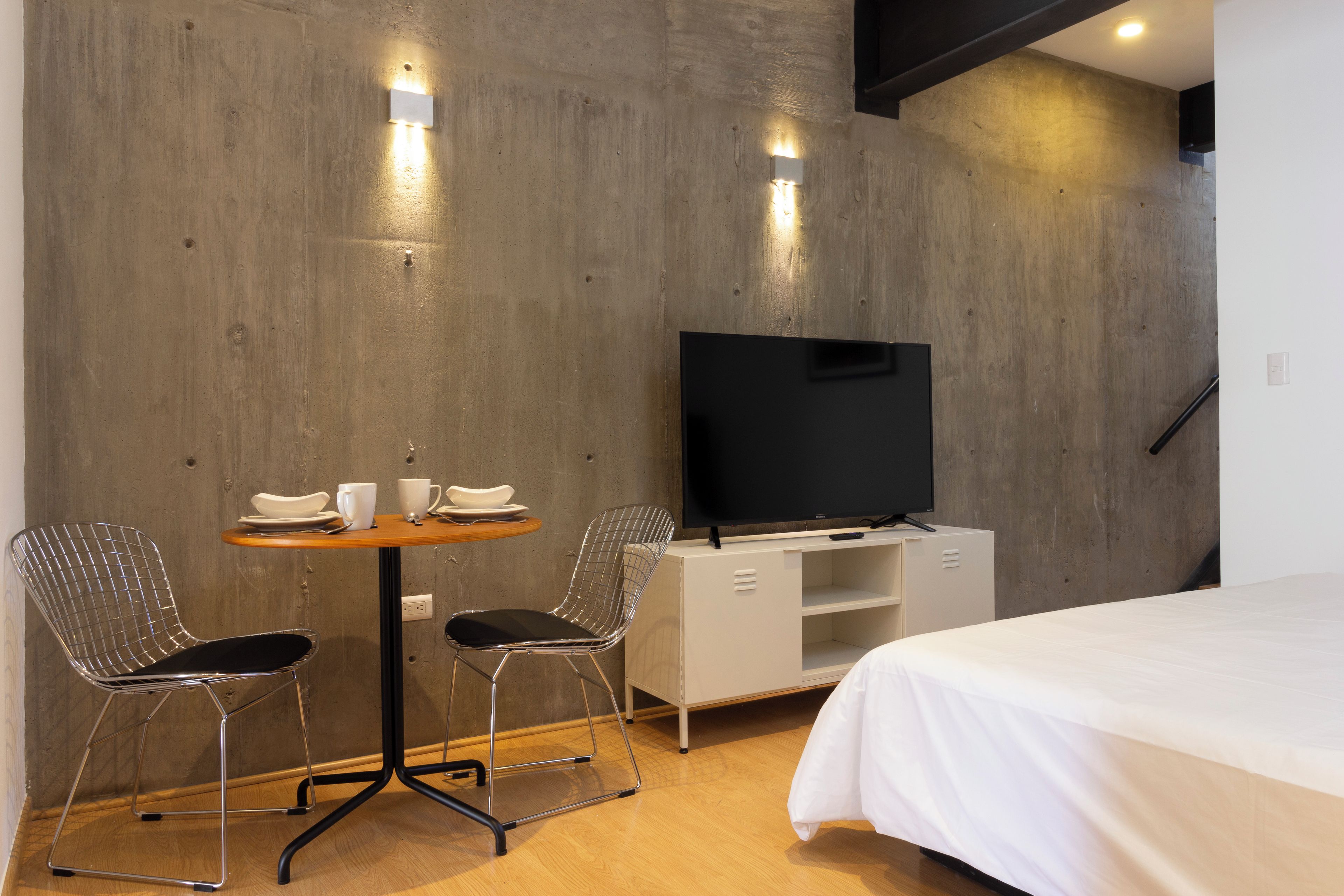 Una habitación de hotel moderna con una cama grande, un televisor de pantalla plana sobre un mueble blanco, una mesita redonda con dos sillas y suelo de madera.