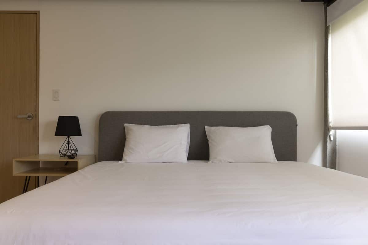 Uma cama bem arrumada com duas almofadas, uma mesa de cabeceira com abajur e uma porta à esquerda.
