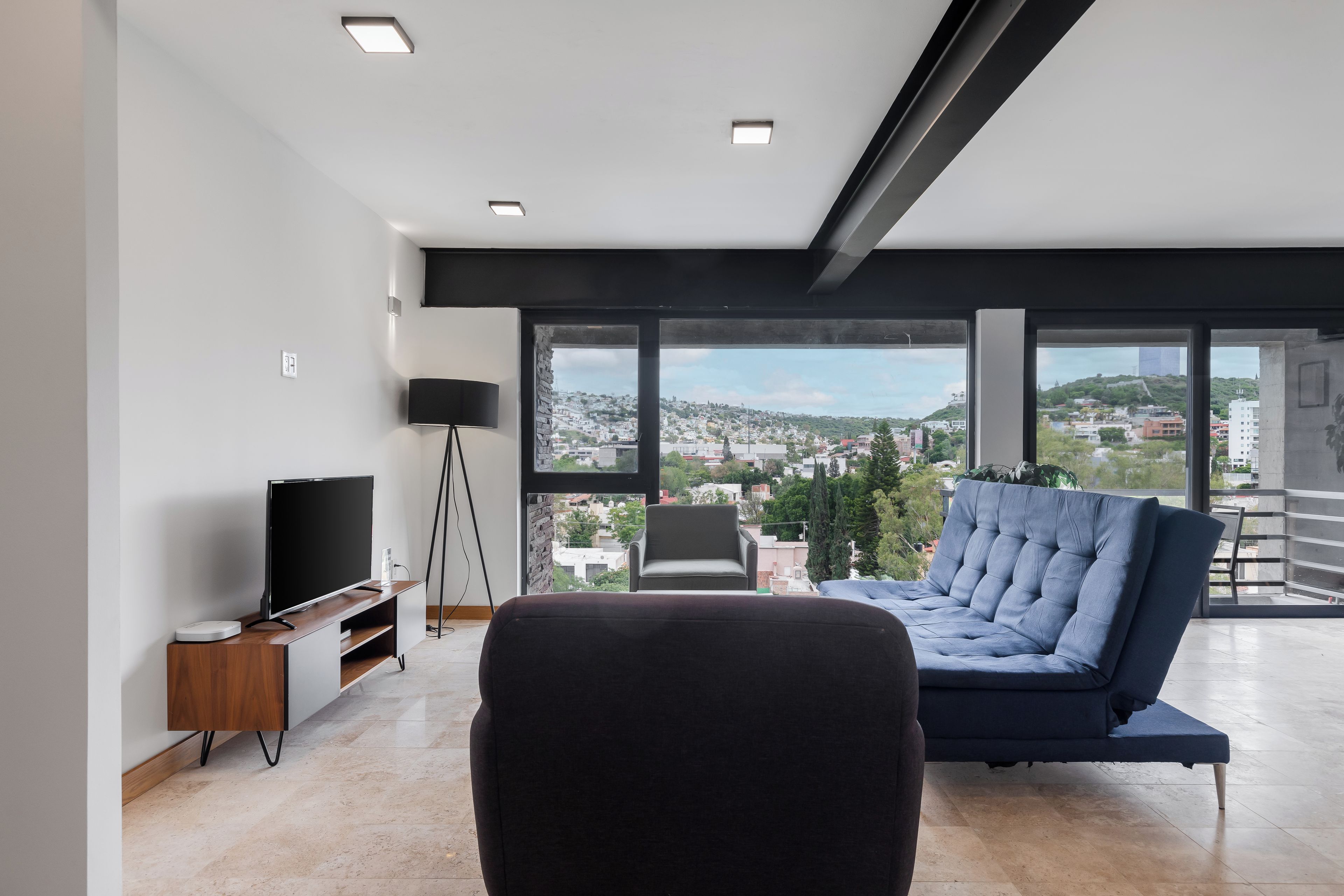 A modern living room with a flat-screen TV, a sofa, and a view of a cityscape through large windows.