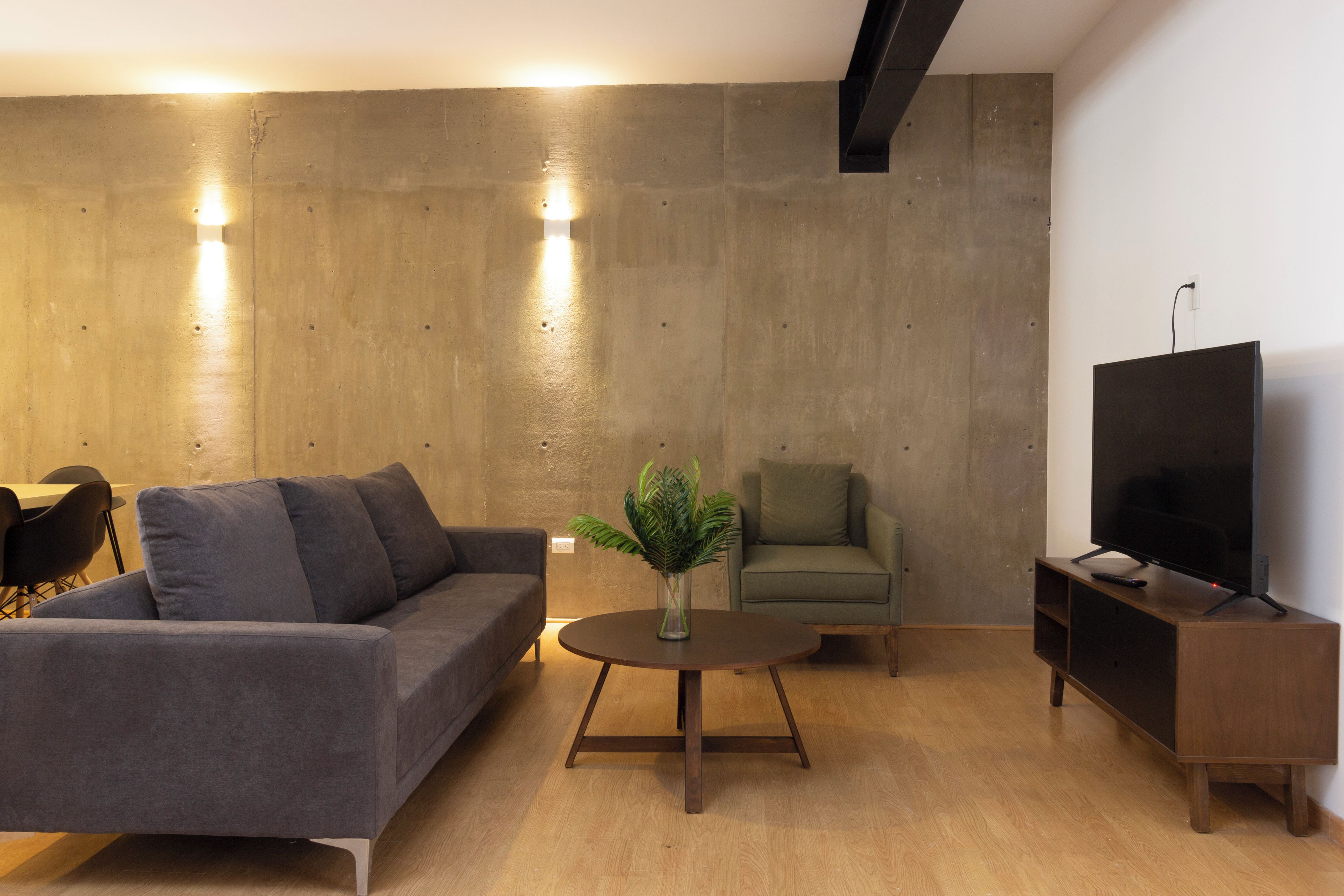 A modern living room with a grey sofa, a round wooden coffee table, a green armchair, a television on a wooden stand, and a concrete accent wall with recessed lighting.