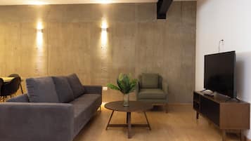 A modern living room with a grey sofa, a round wooden coffee table, a green armchair, a television on a wooden stand, and a concrete accent wall with recessed lighting.