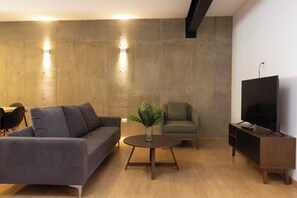 A modern living room with a grey sofa, a round wooden coffee table, a green armchair, a television on a wooden stand, and a concrete accent wall with recessed lighting. - El Doce by Homi Rent (Querétaro)