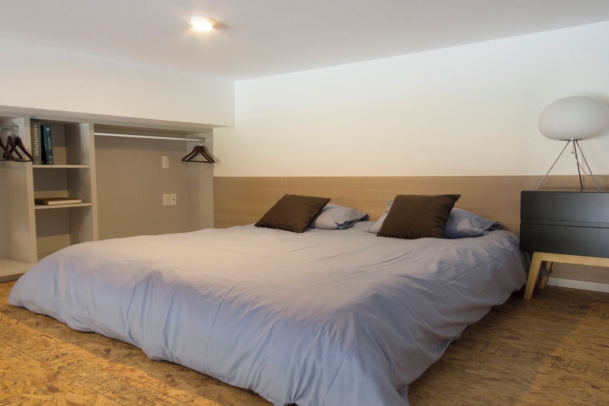 Ett sovrum med en stor säng, en garderob, ett nattduksbord med en lampa och en bokhylla.