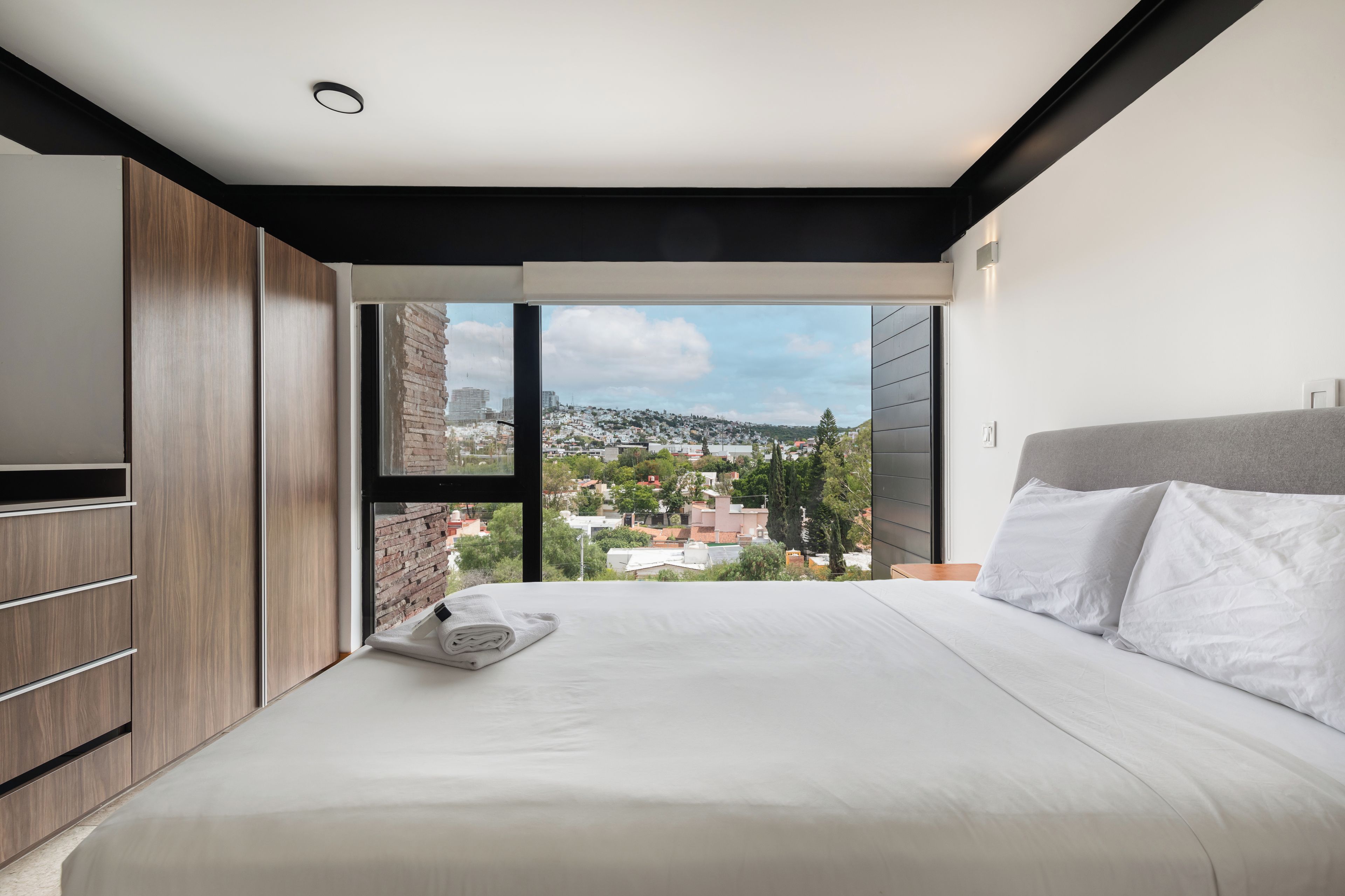 A modern bedroom with a large bed, a wooden wardrobe, and a city view through a large window.