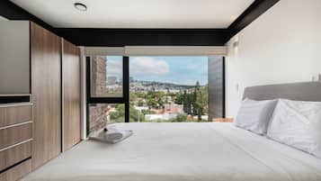 A modern bedroom with a large bed, a wooden wardrobe, and a city view through a large window.