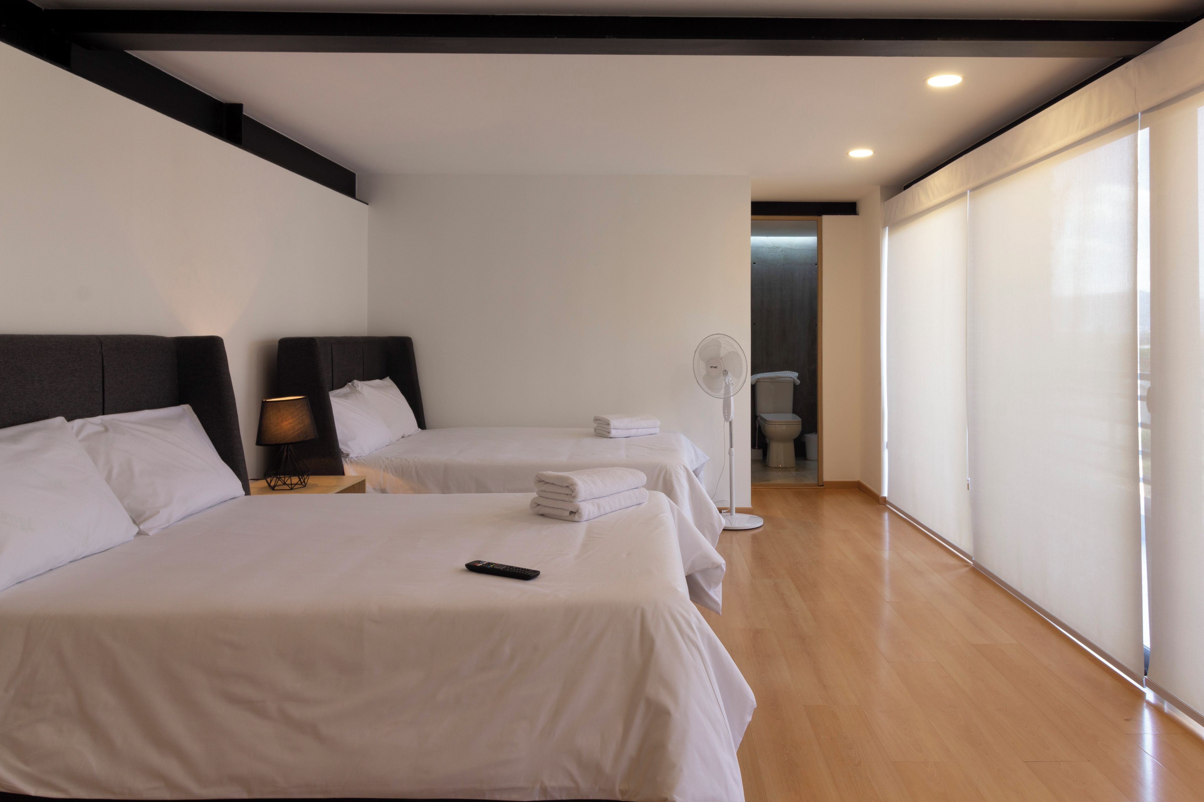 Habitación de hotel con dos camas, una mesita de noche con lámpara, un ventilador y vistas a un pasillo.