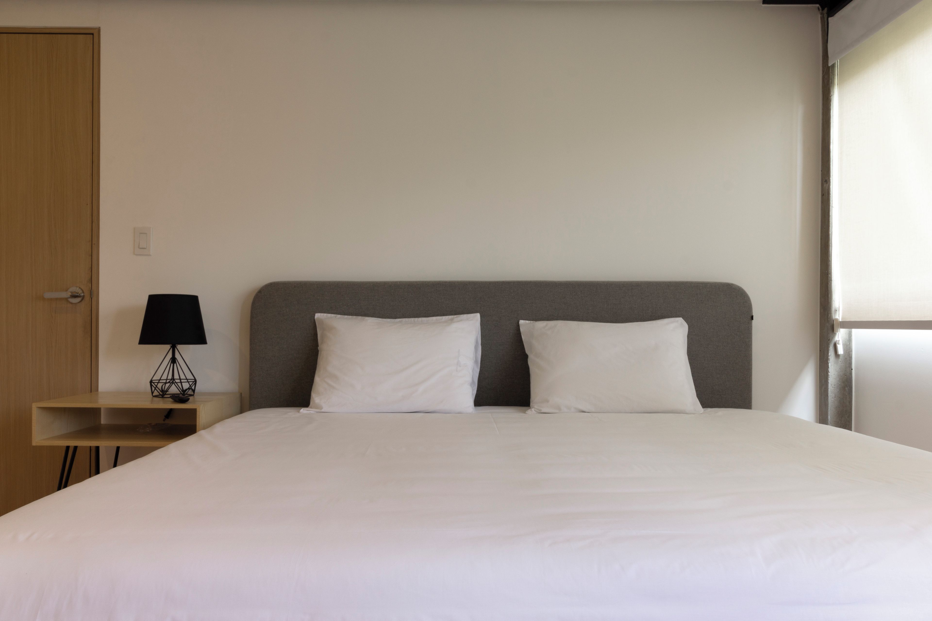A neatly made bed with white linens, a grey headboard, and a small bedside table with a black lamp.