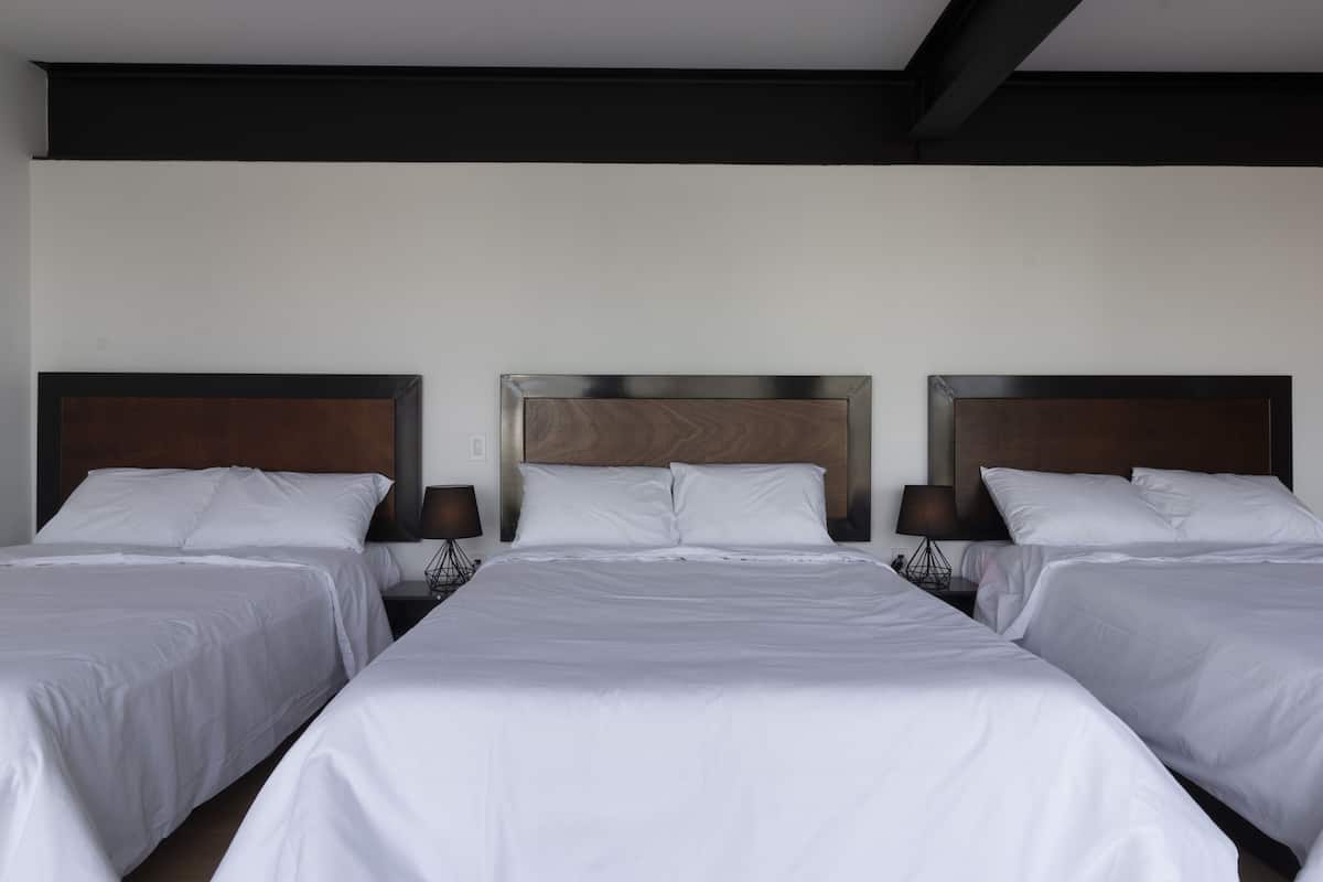 Quarto de hotel com três camas, cada uma com roupa de cama branca e cabeceiras de madeira.