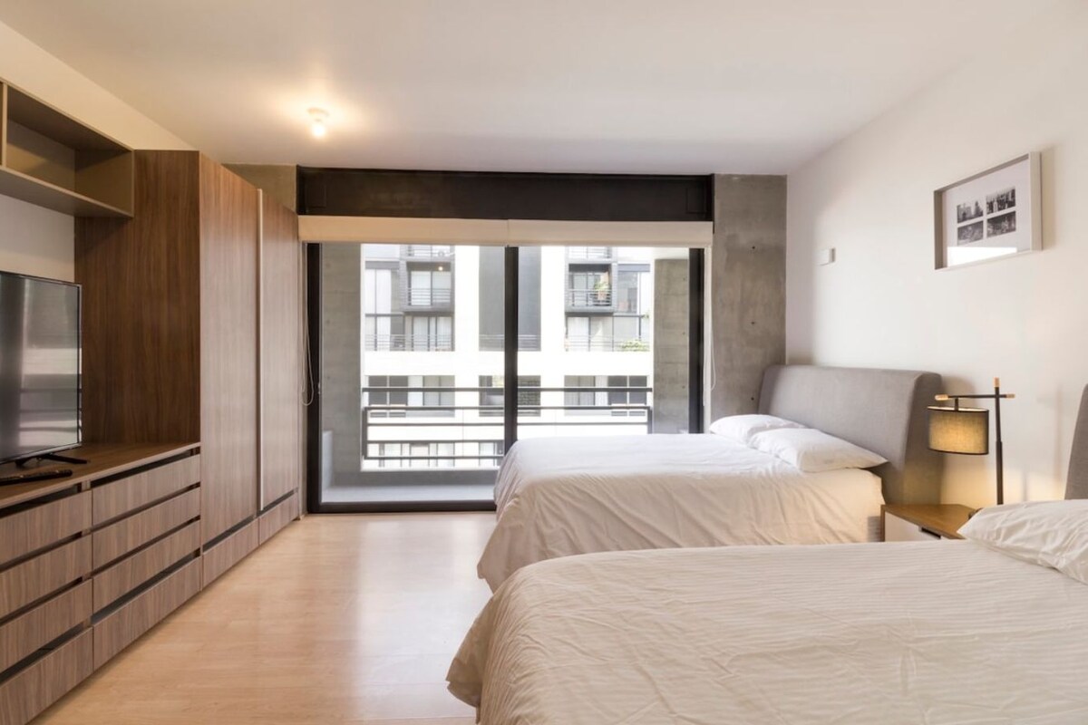A modern bedroom with a TV, two beds, a desk, and a balcony with a city view.