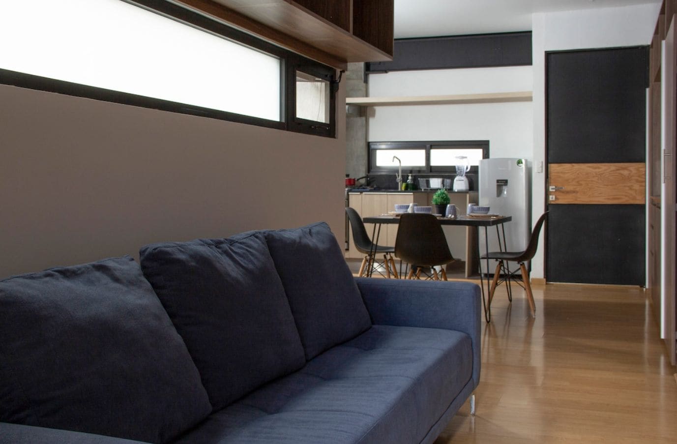 Una sala moderna con un sofá azul oscuro, un comedor con mesa y sillas, y una cocina al fondo.