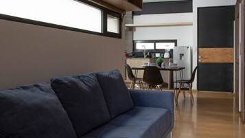 Una sala moderna con un sofá azul oscuro, un comedor con mesa y sillas, y una cocina al fondo.