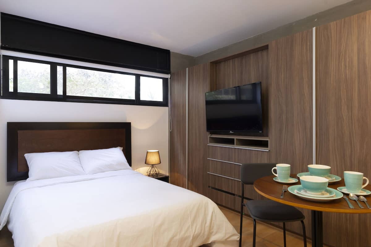 Quarto de hotel com cama, televisão, mesa de jantar com copos e talheres, e janela com persianas.