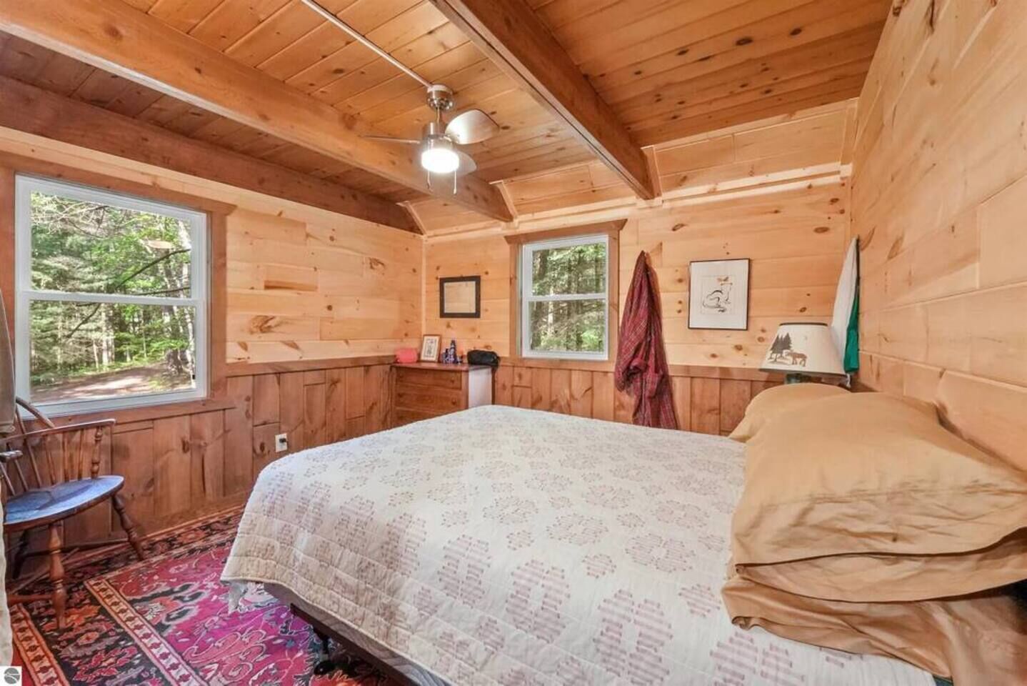 Ein Schlafzimmer einer Holzhütte mit einem Bett, einem Sessel, einem Fenster und einem Deckenventilator.