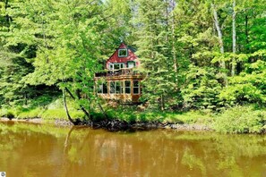 A houseboat with a red roof and multiple decks, surrounded by dense forest and a body of water. - Secluded Riverfront Cottage | Sauna, Fire & Kayaks (Evart)