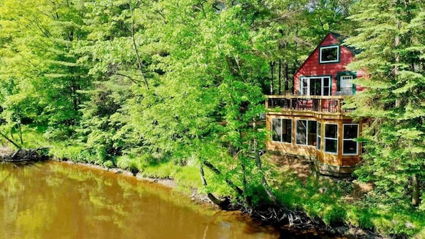 A cabin with a wooden deck and a second-story balcony overlooking a river, surrounded by dense green forest.