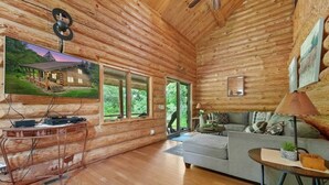 A log cabin interior with a wooden floor, a grey sofa, a TV on a wooden stand, and a large window offering a view of greenery. - Secluded | Sauna, Fire Pit & 20 Acres of Trails (Evart)