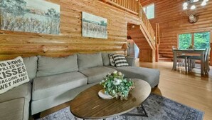 A cozy log cabin interior with a gray sofa, a wooden coffee table with a floral centerpiece, and a dining area with a table and chairs. - Secluded | Sauna, Fire Pit & 20 Acres of Trails (Evart)