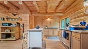 A wooden cabin interior with a kitchen featuring a central island, wooden cabinets, and a stove. - Secluded | Sauna, Fire Pit & 20 Acres of Trails (Evart)