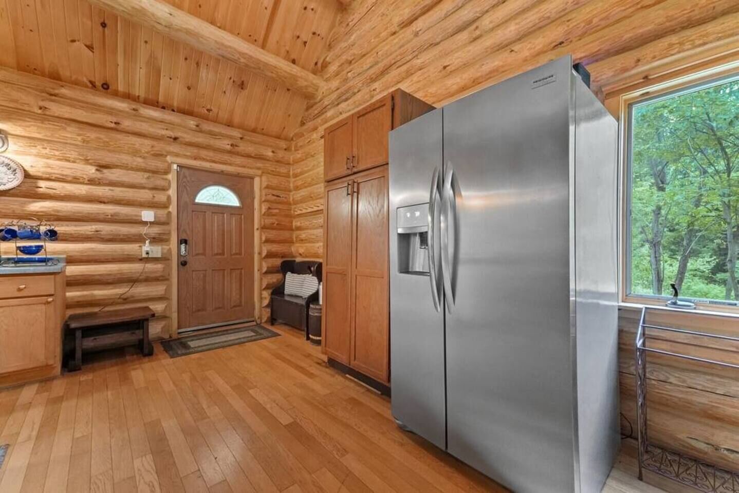 Eine Küche in einer Blockhütte mit Holzwänden, einem Edelstahlkühlschrank, Holzschränken, einer Bank und einem Fenster mit Blick auf Bäume.
