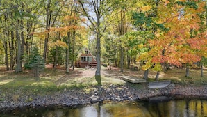 A cabin surrounded by trees with autumn foliage, a small dock, and a pond. - A-Frame | Hot Tub, Fireplace, River, Kayaks &Tubes (Sears)