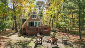 A cabin with a deck, surrounded by trees and a fire pit area with chairs. - A-Frame | Hot Tub, Fireplace, River, Kayaks &Tubes (Sears)