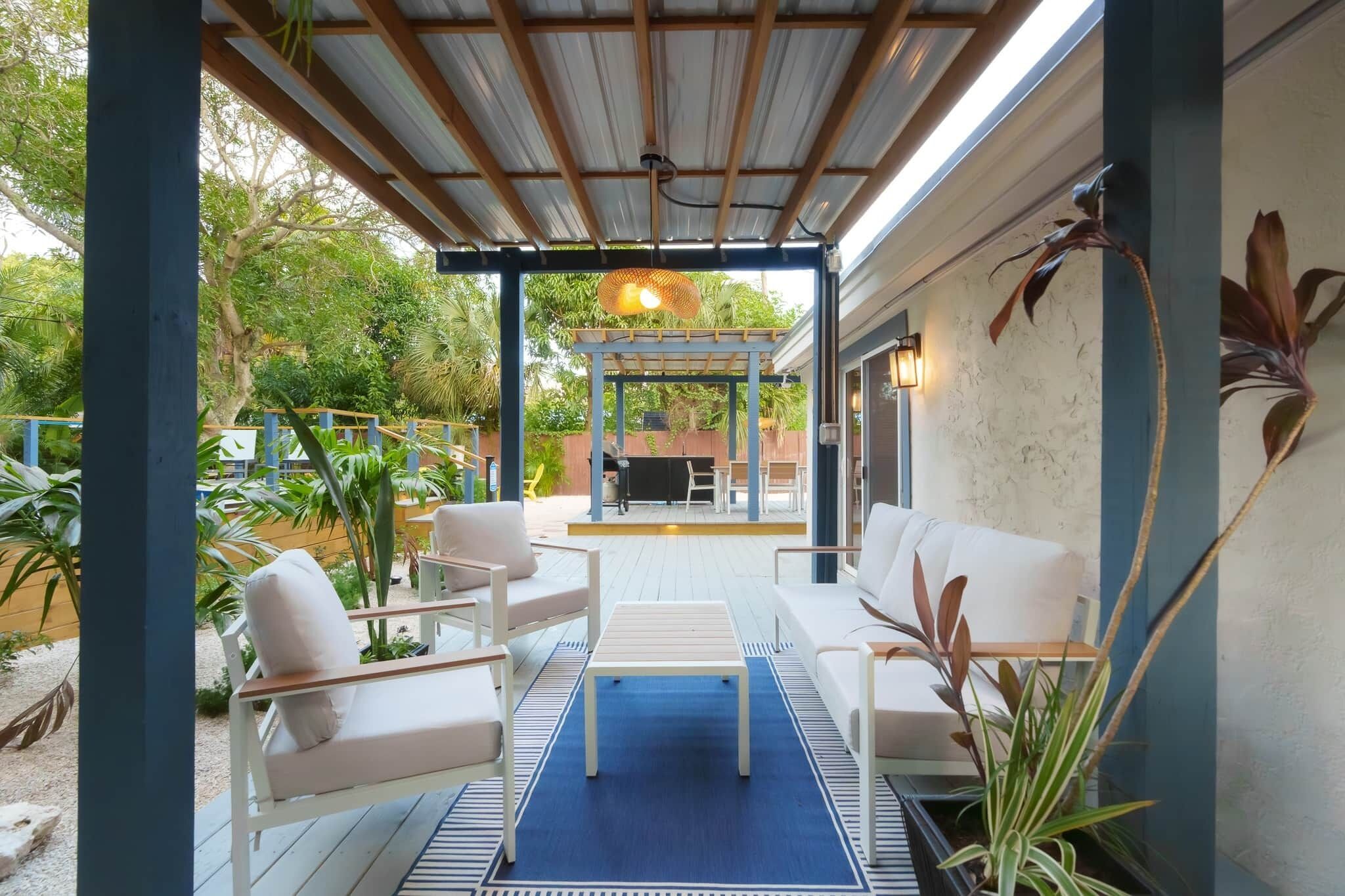 Un patio exterior con mobiliario blanco, un tapete azul y una pérgola con una lámpara colgante.