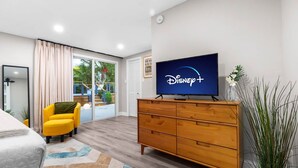 Ein modernes Hotelzimmer mit einem Bett, einem gelben Sessel, einer Holzkombination mit einem Fernseher, der das Disney+-Logo zeigt, einem großen Fenster mit Vorhängen und Blick ins Freie.