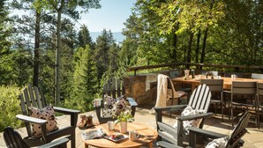 An outdoor seating area with Adirondack chairs and a wooden table, surrounded by a stone wall and a view of a forested landscape. - Outpost: Granite Ridge Chateau (Teton Village)