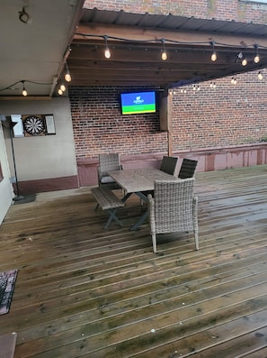 Eine Terrasse im Freien mit einem Holztisch, Korbstühlen, einem Dartspiel und einem an einer Ziegelwand montierten Fernseher.