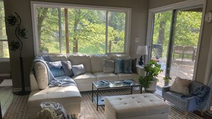 A living room with a sectional sofa, a coffee table, and a small side table. There is a large window providing a view of trees and a patio area. - Lakefront cottage on rustic setting in cove (Buckhead)