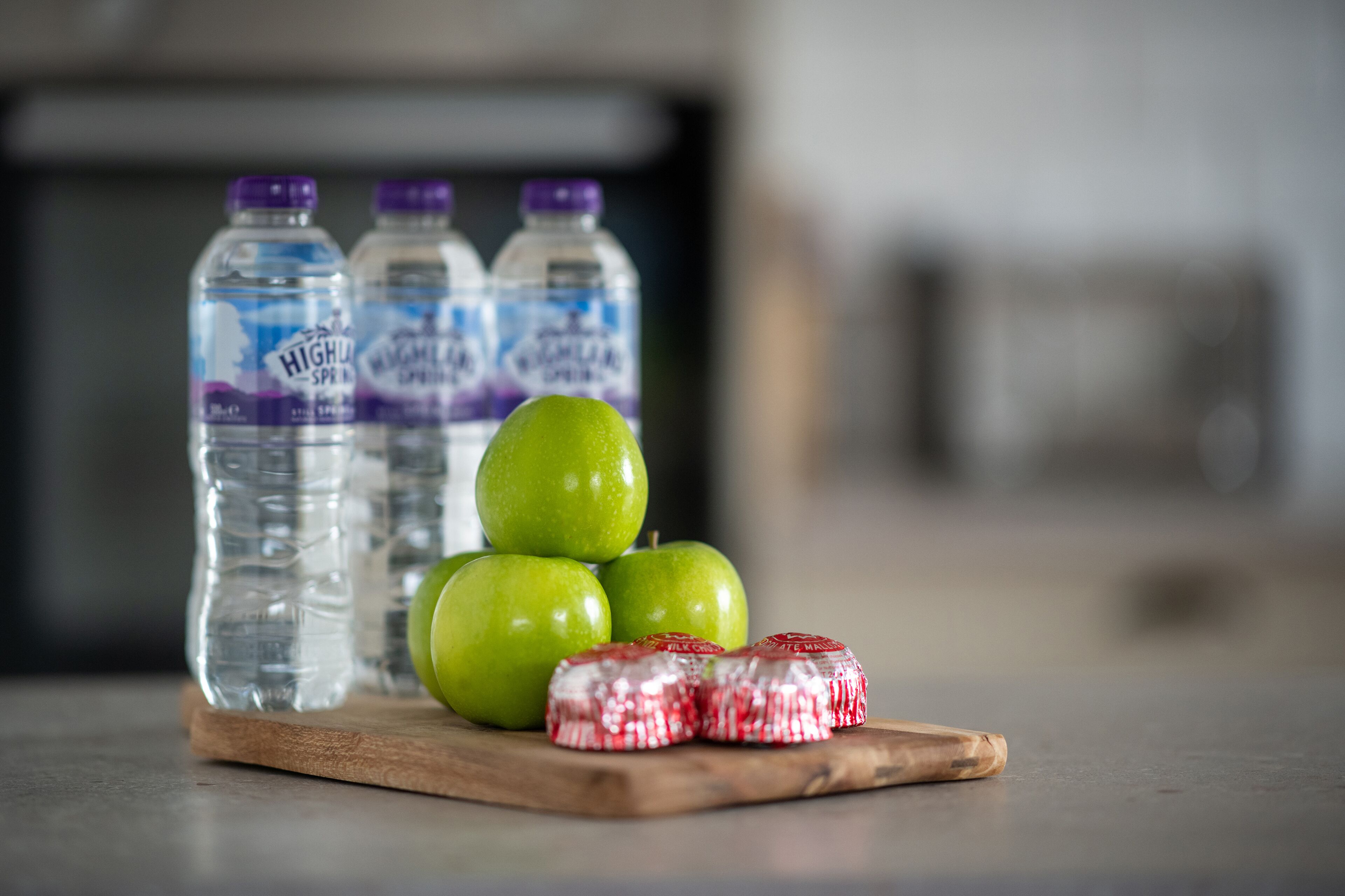 A wooden cutting board with green apples and red and white candy bars, with water bottles in the background.