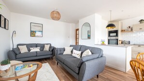 Ein modernes Wohnzimmer mit einem grauen Sofa, einem Glastisch und einem kleinen Esstisch. Es gibt eine Küchenzeile mit weißen Schränken und einem schwarzen Ofen.