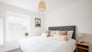 Ein ordentlich eingerichtetes Schlafzimmer mit einem großen Bett, einem Nachttisch mit Lampe, einem kleinen Topfpflanzen und einem Fenster mit Jalousien.