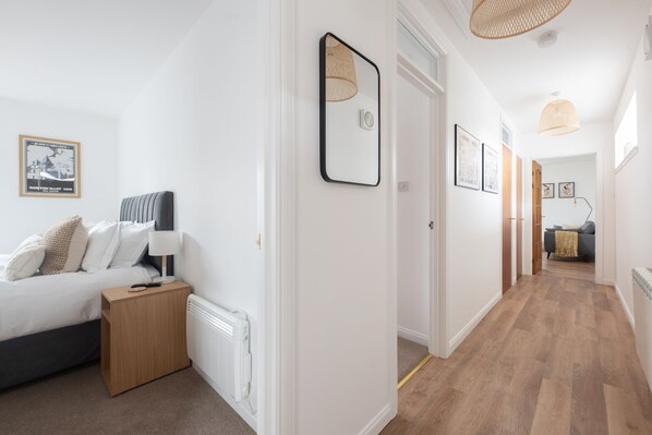 Un couloir d’hôtel avec un lit, une table de chevet et une lampe sur le côté gauche. Les murs sont ornés de tableaux encadrés et un luminaire est suspendu au plafond.