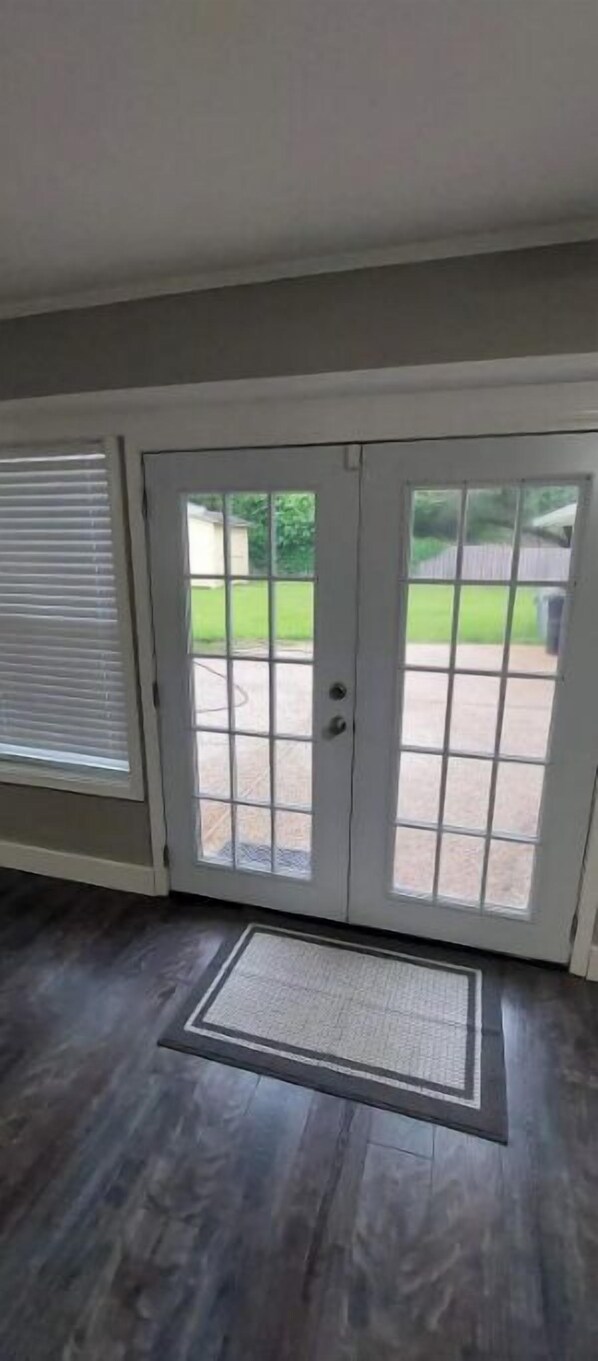 A room with a dark wooden floor, a white door with glass panels, and a view of a green lawn and a building in the background. - 4 B/R HOUSE. Central Location. Clean and comfy. (Baton Rouge)