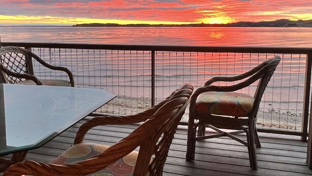 A deck with wooden chairs and a table, overlooking a sunset over water. - Right on the Lake Michigan - walk to town and across from TART trail (Suttons Bay)