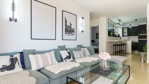 Een moderne woonkamer met een glazen salontafel, een grijze bank met gekleurde kussens en kunstwerken in lijsten aan de muur.
