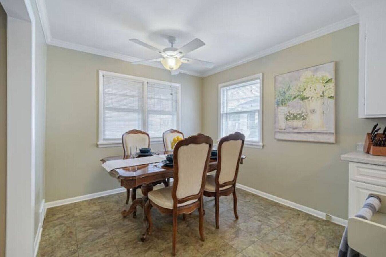 Uma sala de jantar com uma mesa de madeira e quatro cadeiras, um ventilador de teto e um quadro na parede.