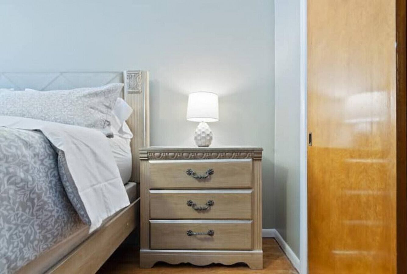 Quarto com cama, mesa de cabeceira e abajur, e porta de madeira.