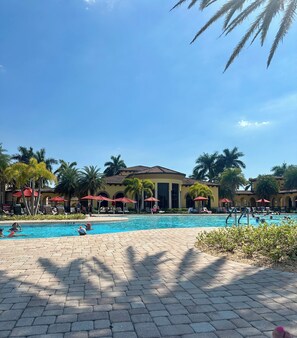 Área de piscina com espreguiçadeiras e guarda-sóis, rodeada por palmeiras e um edifício com arcos.