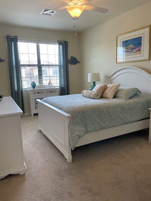 A bedroom with a bed, bedside tables, a nightstand, a ceiling fan, a window with curtains, and a wall-mounted artwork.