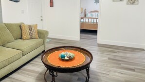Una sala de estar con un sofá verde, una mesa de centro redonda de madera con adornos, una puerta blanca y un pasillo que conduce a un dormitorio.