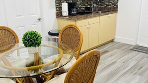 Cocina compacta con cafetera, tostadora y lavamanos. Zona de comedor con mesa de vidrio y sillas de mimbre.
