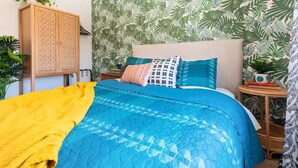 Un dormitorio con una cama, un armario de madera, un muro con estampado de hojas tropicales y una mesita con libros y una planta.