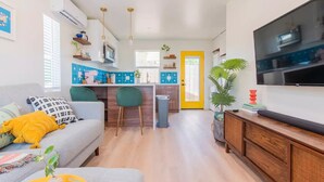 Una sala de estar moderna con un sofá, sillas verdes, un televisor y una zona de cocina con una puerta amarilla.